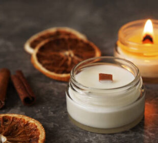 Handmade burning candle with wooden wick in white glass jar with cinnamon and dry oranges at the background, winter cozy home decoration