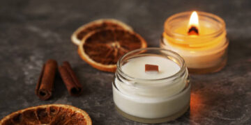 Handmade burning candle with wooden wick in white glass jar with cinnamon and dry oranges at the background, winter cozy home decoration