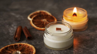Handmade burning candle with wooden wick in white glass jar with cinnamon and dry oranges at the background, winter cozy home decoration