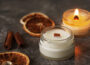 Handmade burning candle with wooden wick in white glass jar with cinnamon and dry oranges at the background, winter cozy home decoration